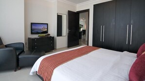 A modern hotel room with a large bed, a TV, a dark wardrobe, and a seating area with a chair and a table. - Retaj Residence Al Corniche (Doha)