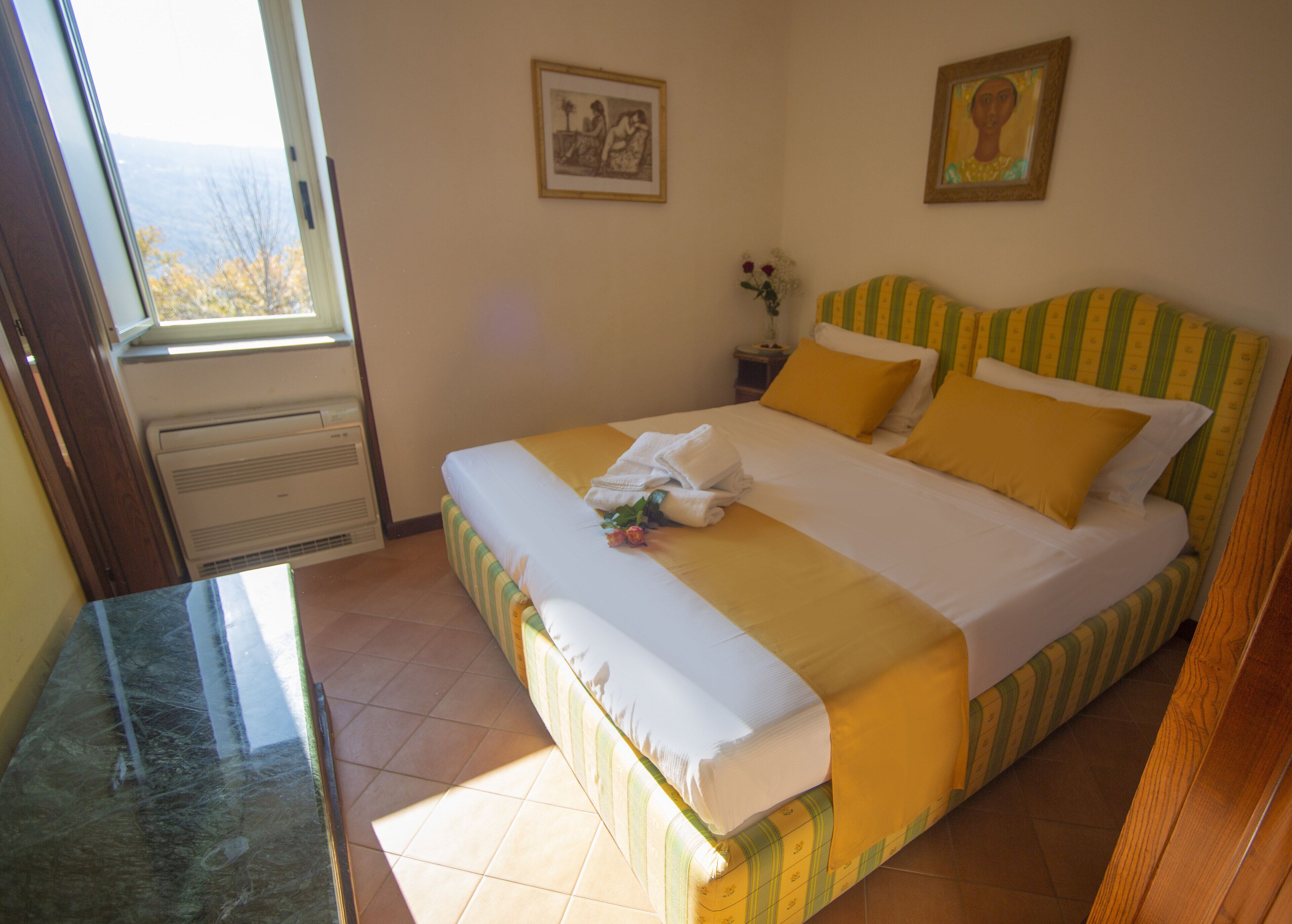 A neatly made bed with yellow and white bedding, a window with a view of trees, and two framed pictures on the wall.
