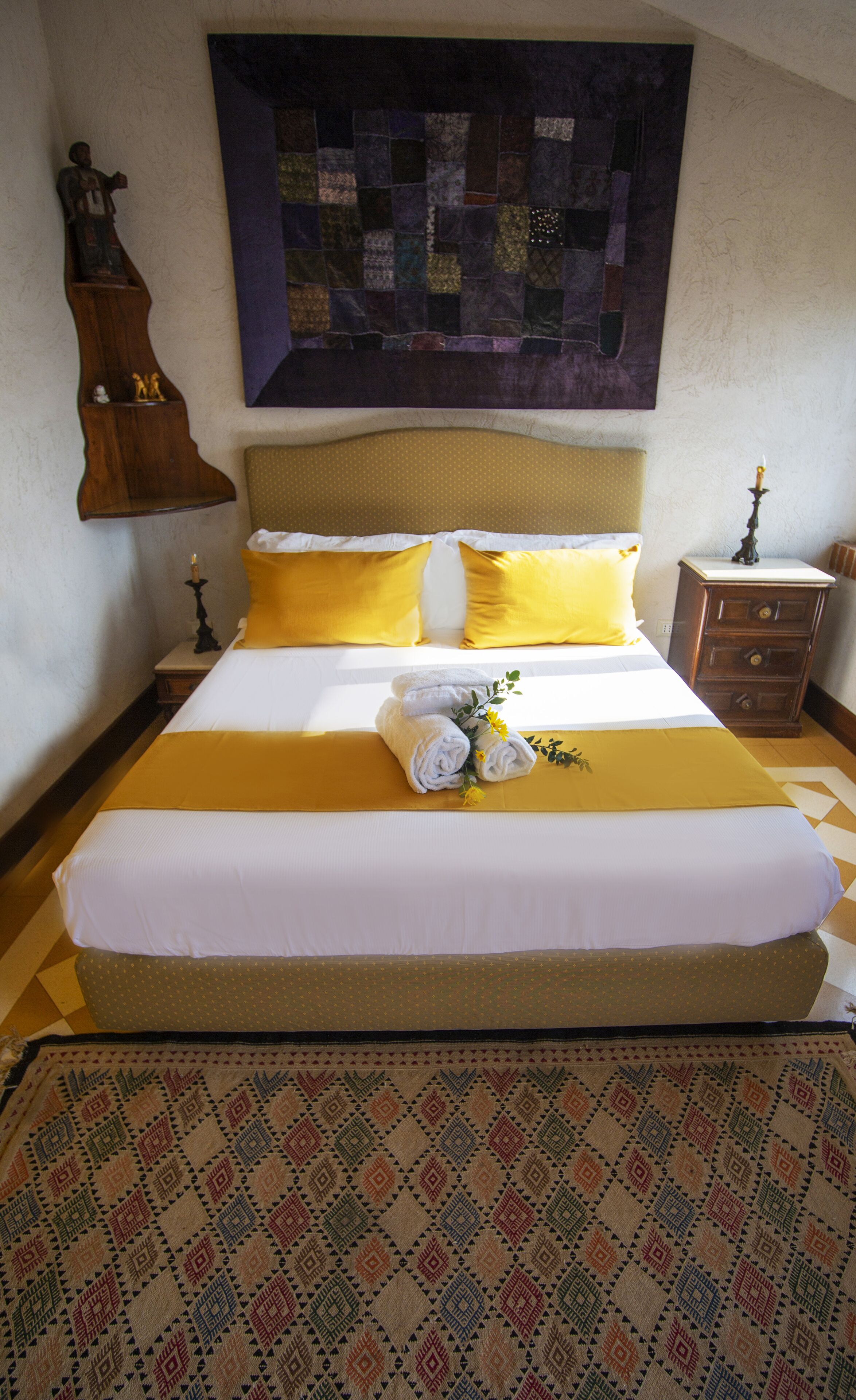 Una cama bien hecha con ropa de cama amarilla y blanca, un edredón encima y una alfombra estampada en el suelo.