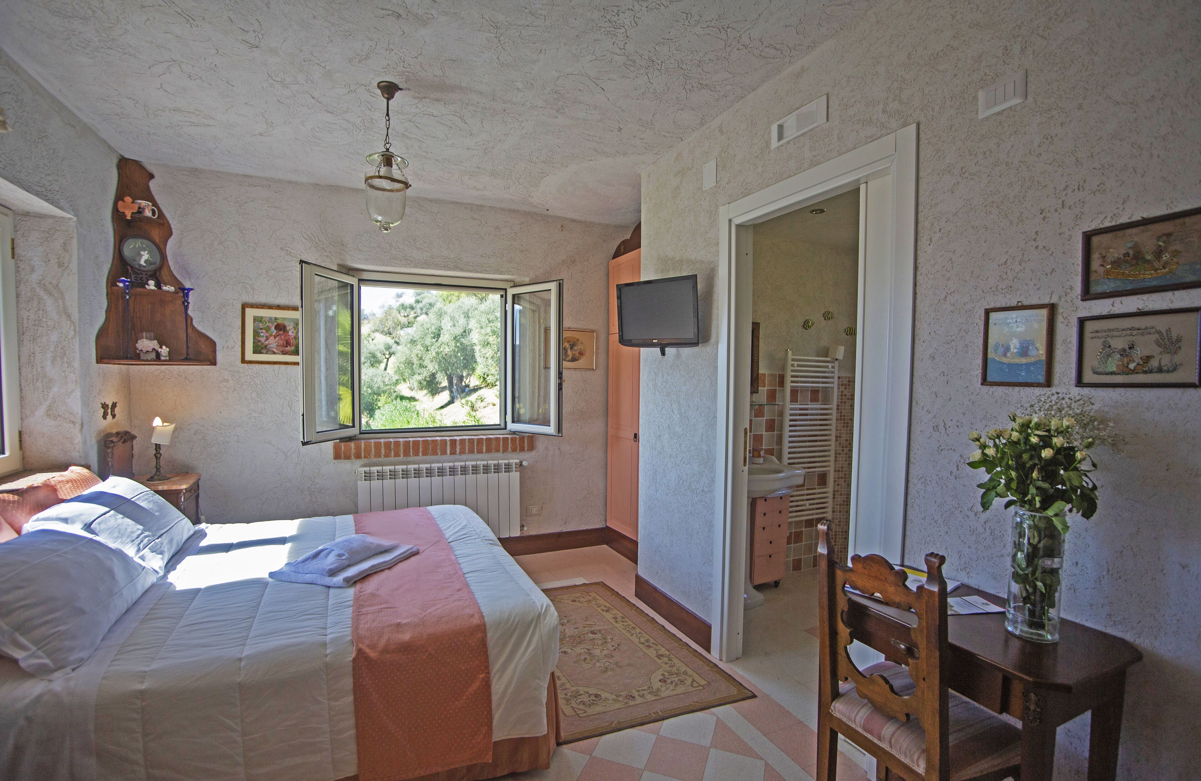 A bedroom with a bed, a window, a television, and a desk with a vase.