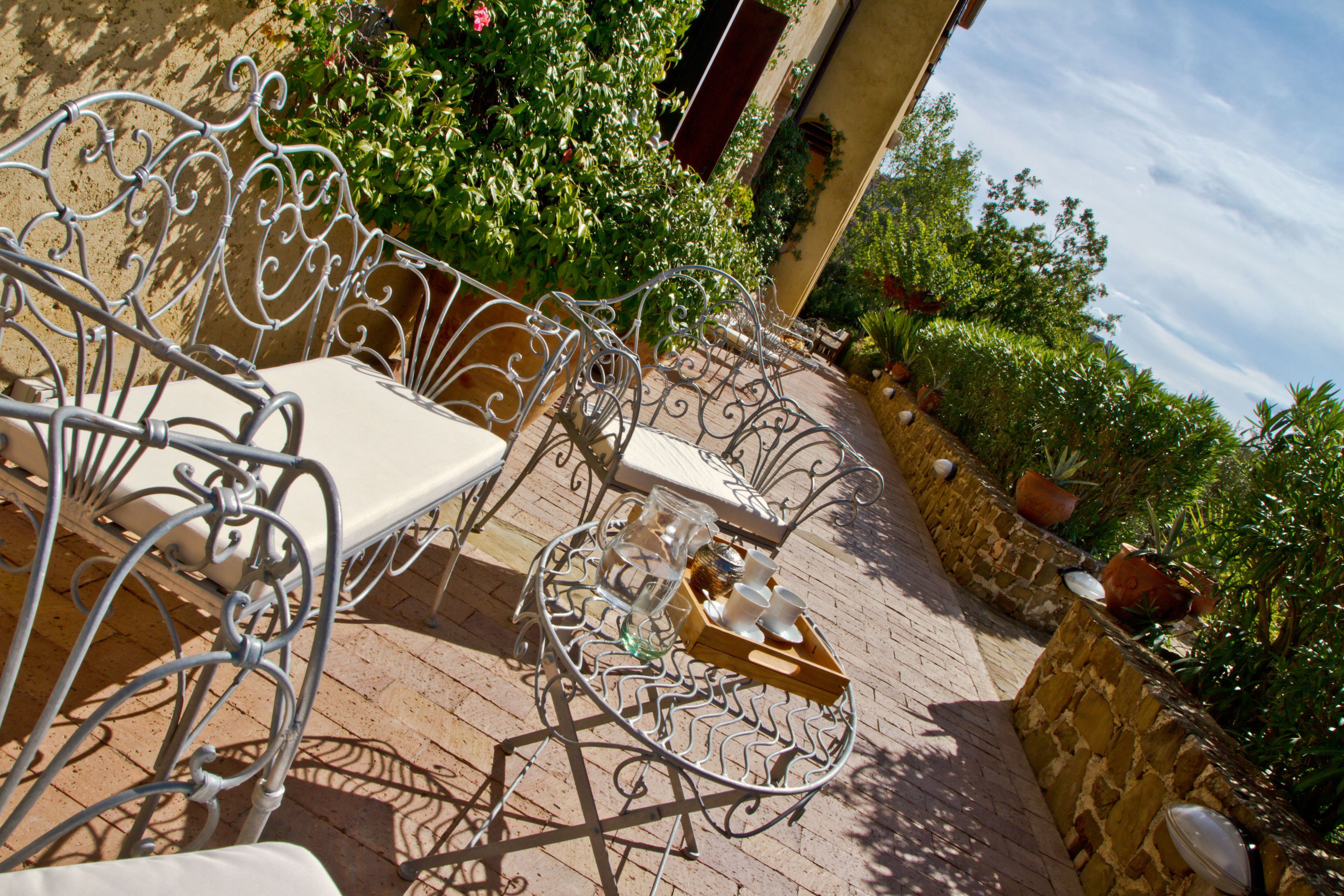 Une terrasse avec des chaises et une table en métal, entourée de plantes en pot et d’un mur de pierre.