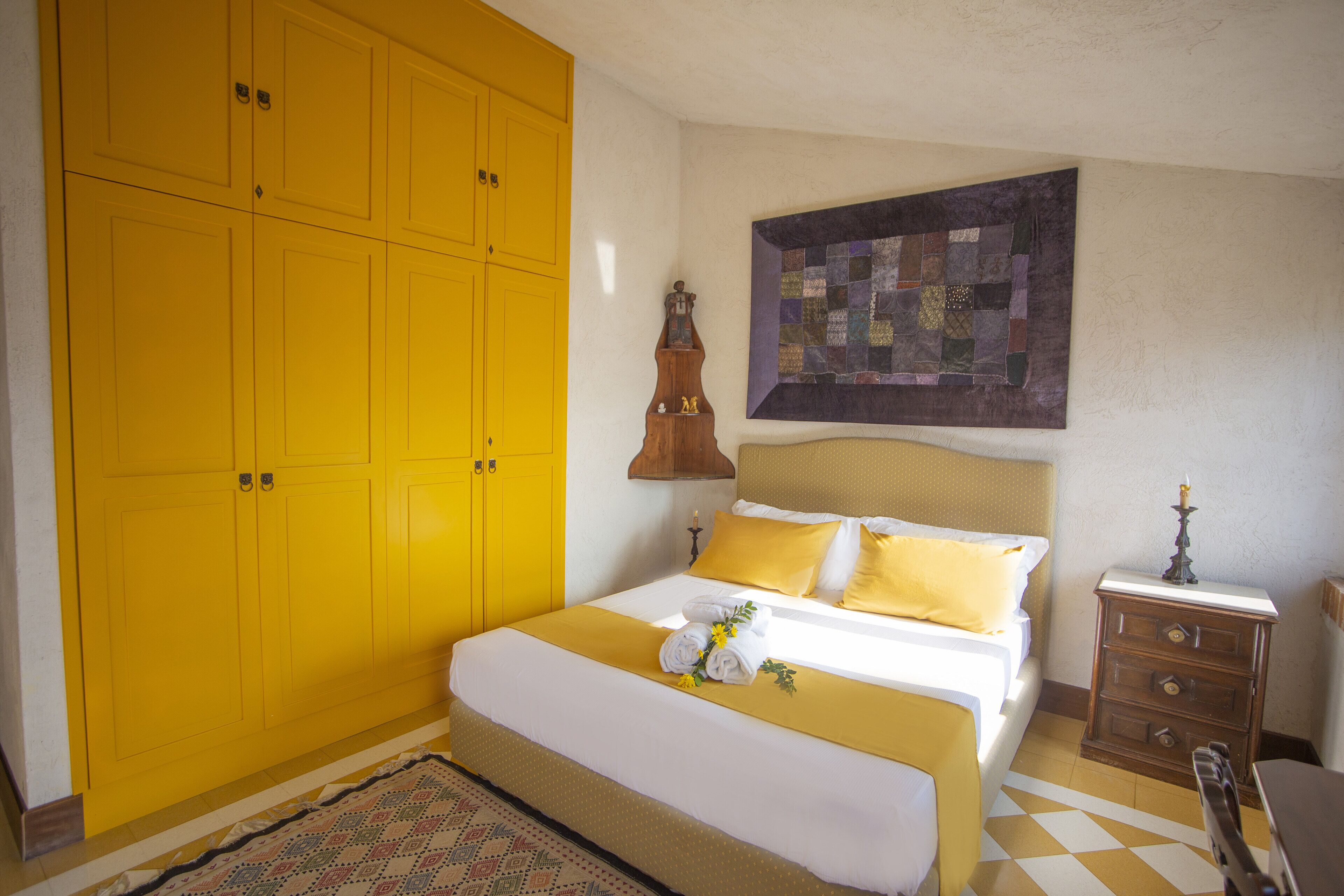 Ein Hotelzimmer mit einem großen, gelben Kleiderschrank, einem Bett mit weißer und gelber Bettwäsche, einem gewölften Wandbild und einem kleinen Nachttisch aus Holz.