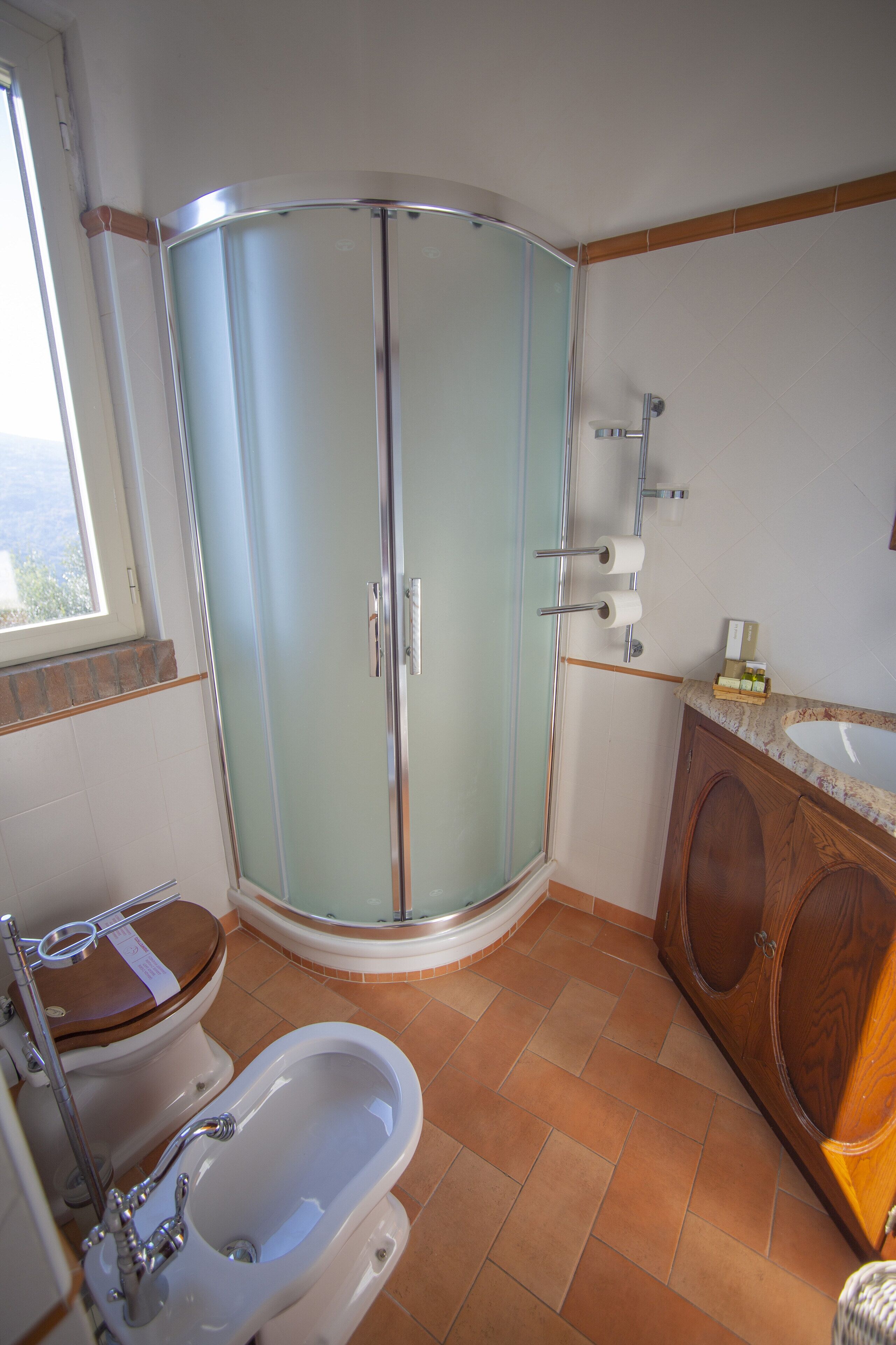 A bathroom with a glass-enclosed shower, a wooden vanity, a white sink, and a toilet with a wooden seat.