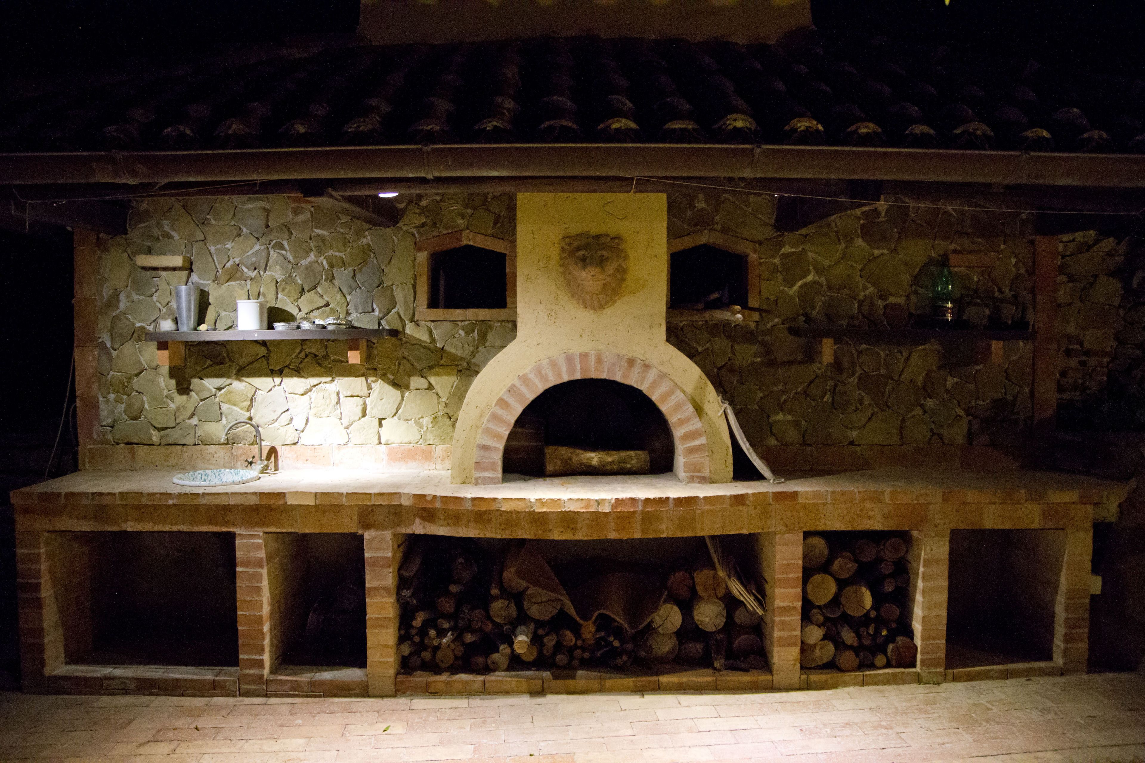 A stone-built outdoor kitchen with a pizza oven, shelves with bottles, and a fire pit area.