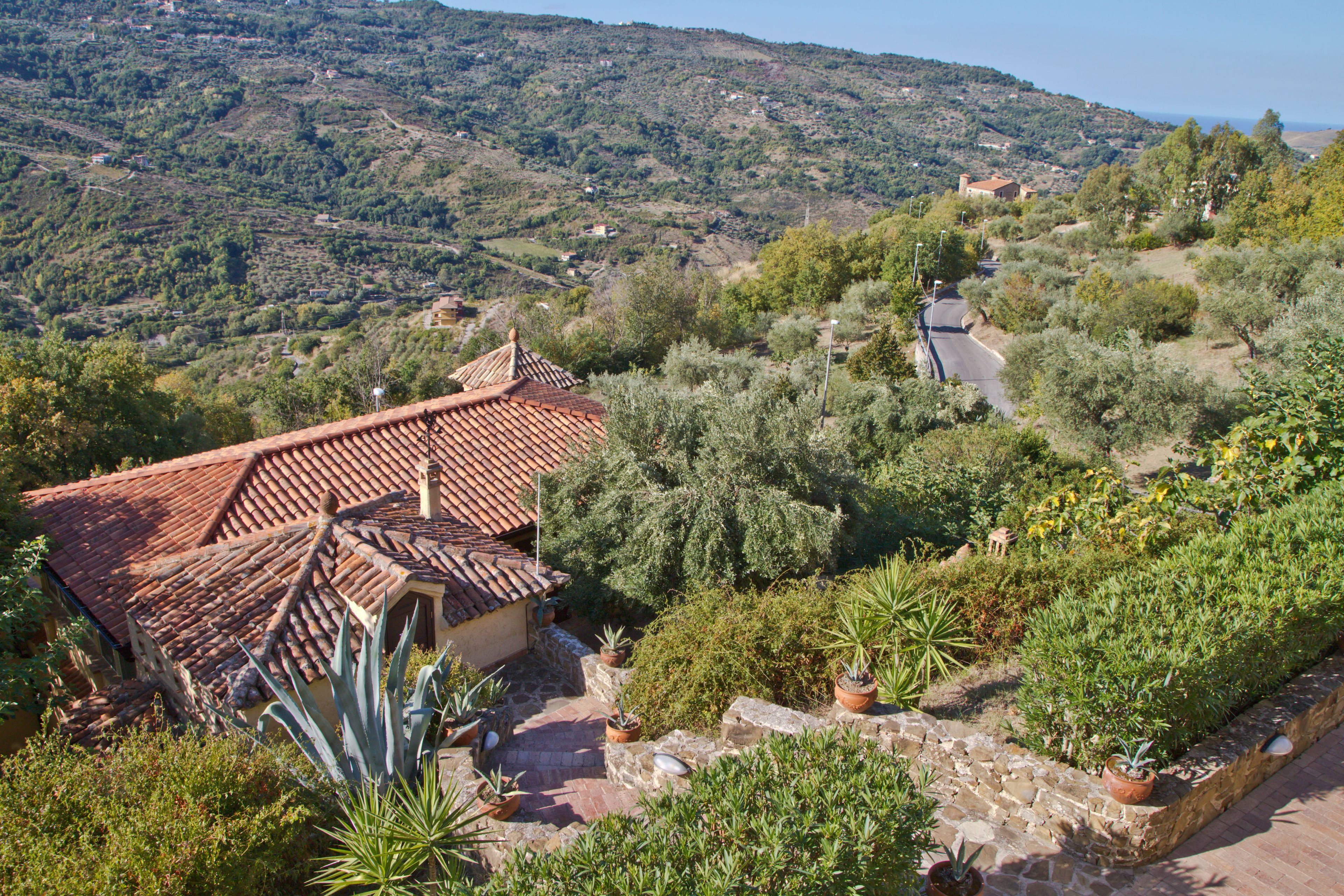 A terraced hillside with a traditional building featuring a red-tiled roof, surrounded by lush greenery and a clear view of the distant landscape.