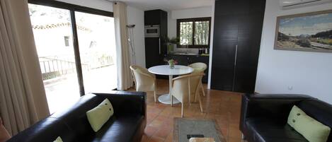 A living room with two black sofas, a coffee table with decorative items, a dining table with chairs, and a kitchen area with a window.