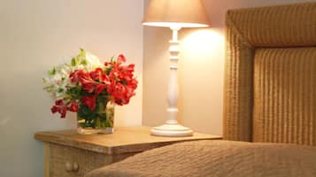 Una cama con una manta color beige, una mesita de noche con una lámpara blanca y un florero con flores rojas y blancas.