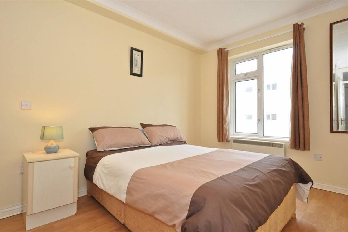 A bedroom with a bed, a nightstand with a lamp, a window with curtains, and a framed picture on the wall.