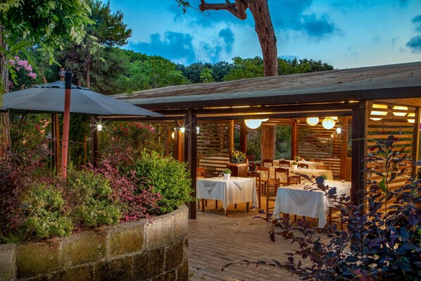 Un área de comedor al aire libre con mesas y sillas, rodeada de plantas y una pérgola de madera.