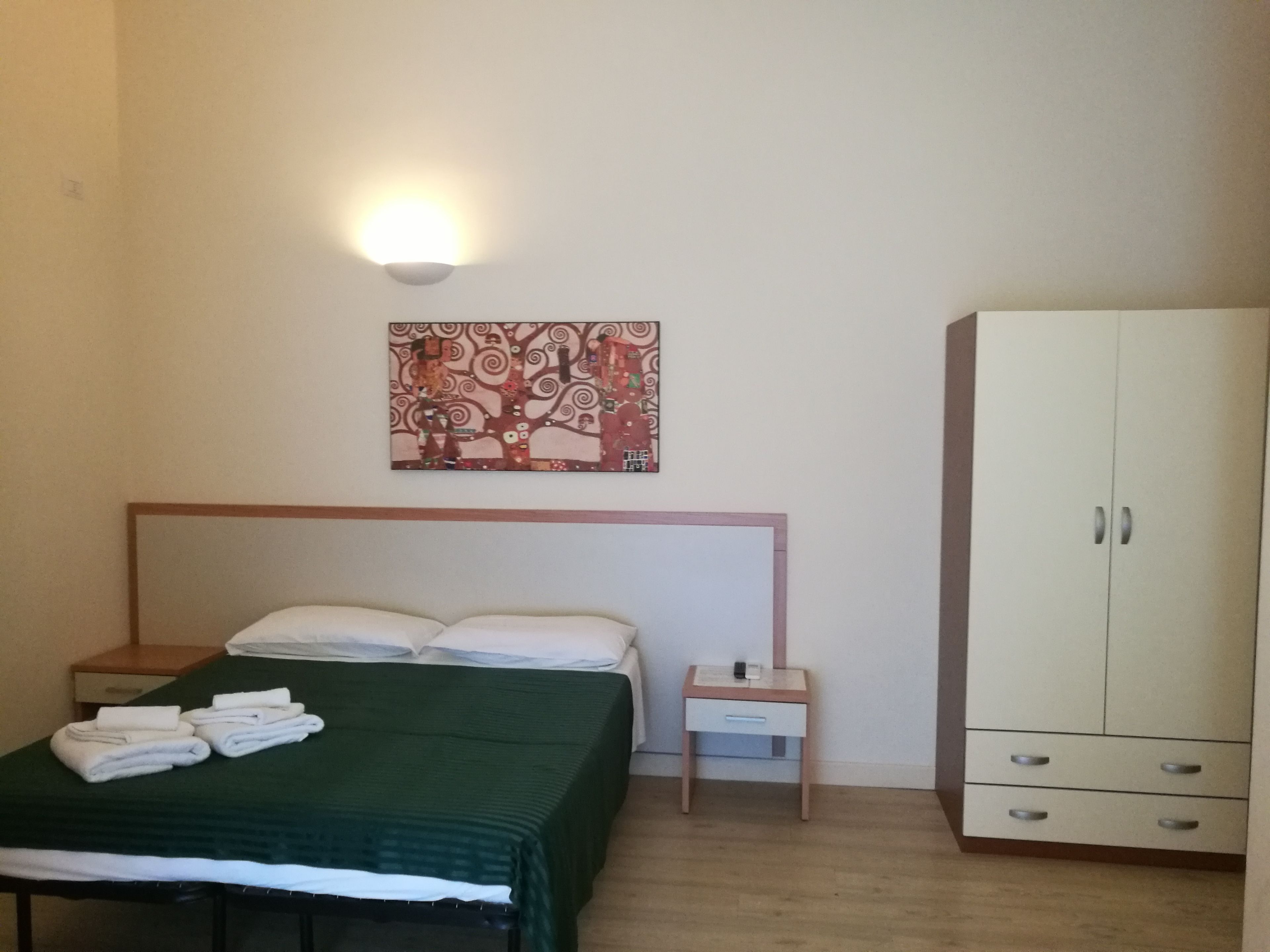 A hotel room with a bed, a nightstand, a small table, a wardrobe, and a wall-mounted light fixture.