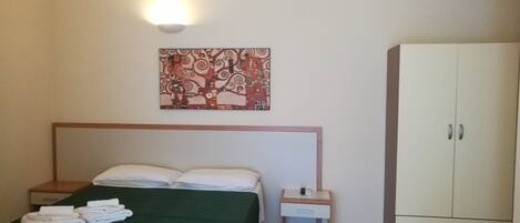 A hotel room with a bed, a nightstand, a small table, a wardrobe, and a wall-mounted light fixture.