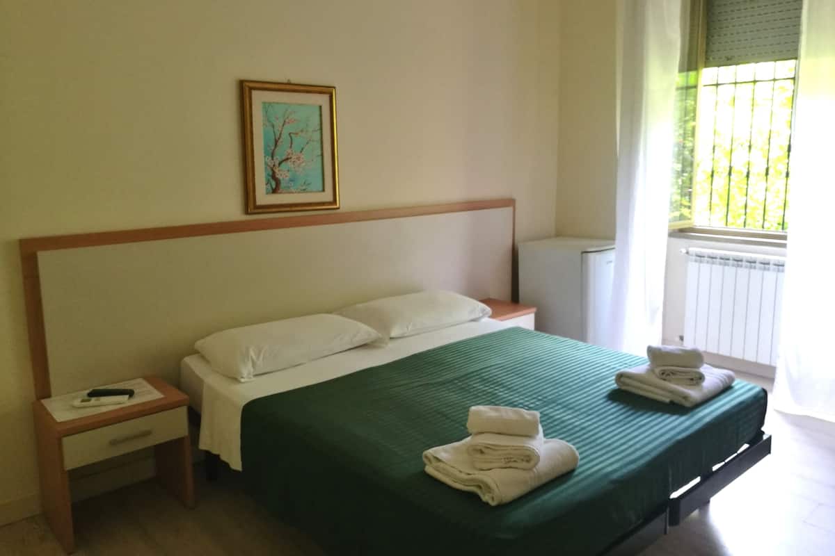 A hotel room with a single bed, a nightstand, a small table, a framed picture on the wall, and a window with a view of greenery.