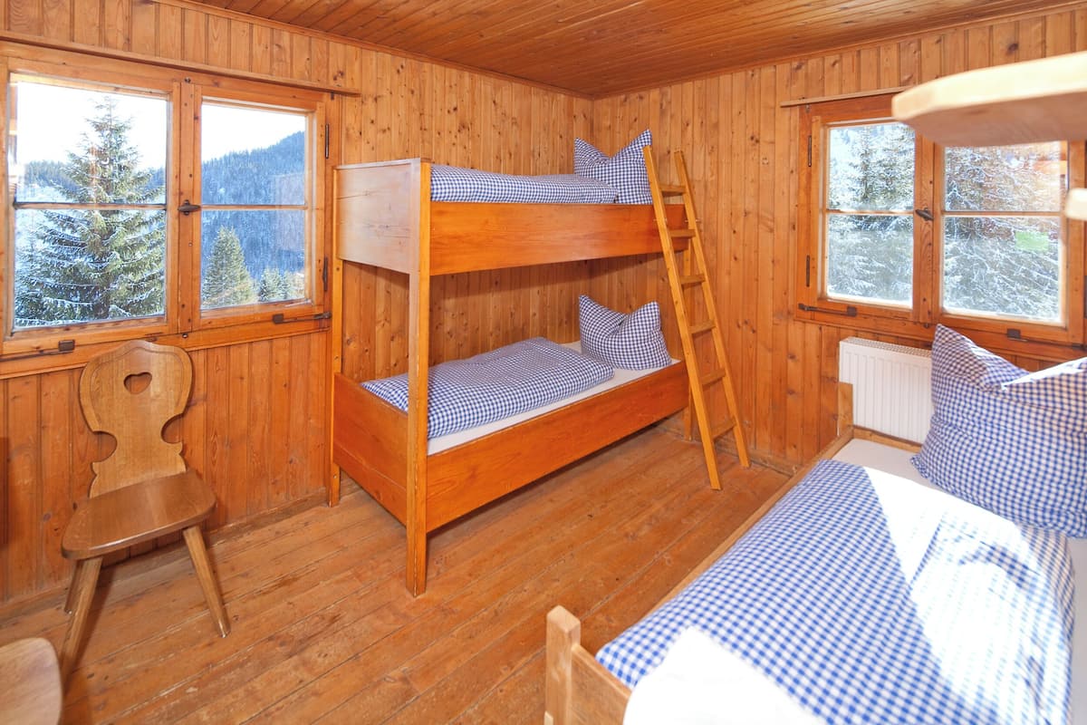 A wooden cabin with bunk beds, a ladder, a chair, and large windows with a forest view.