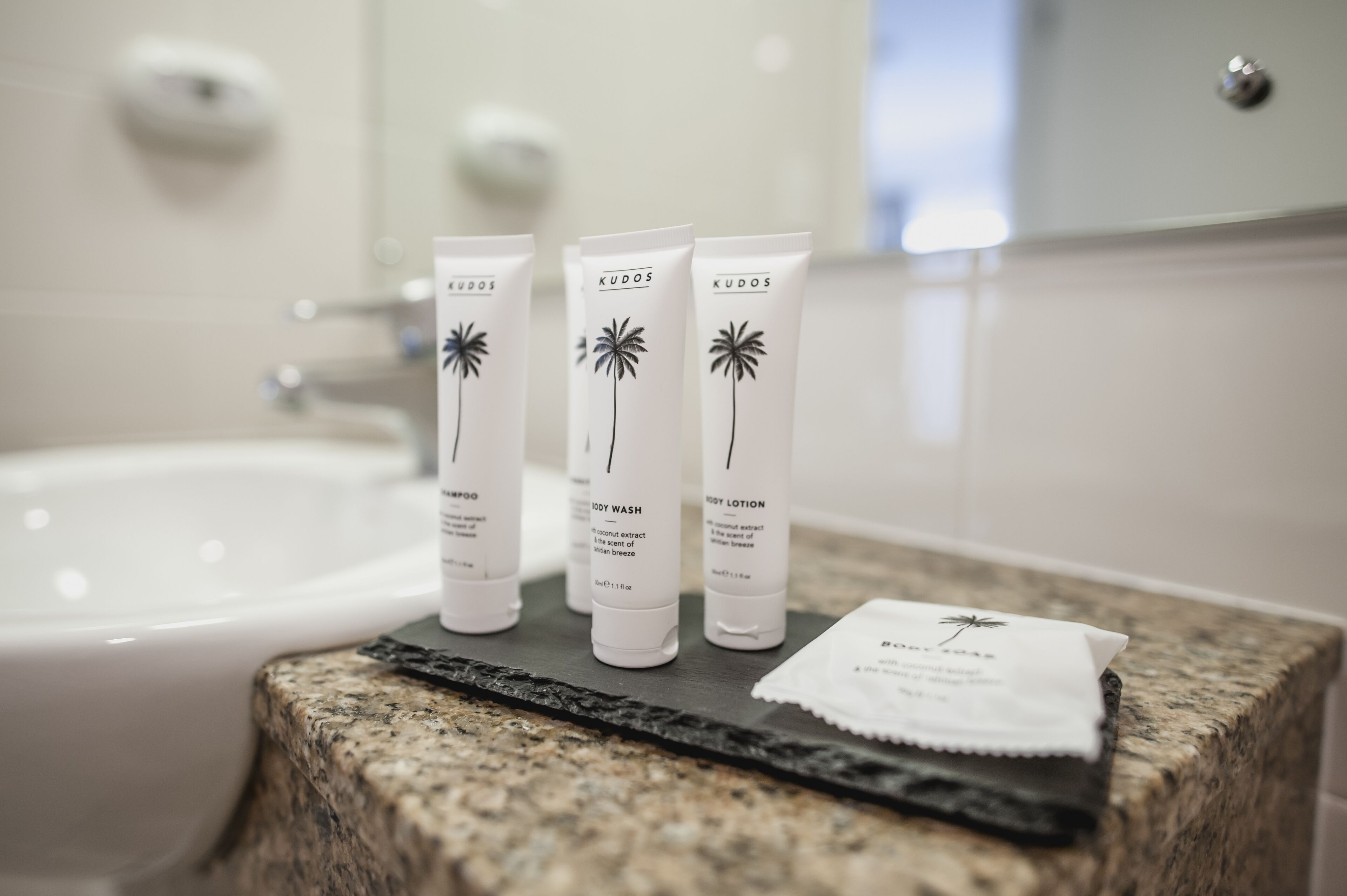 Three KUDOS body care products on a granite countertop in a bathroom.