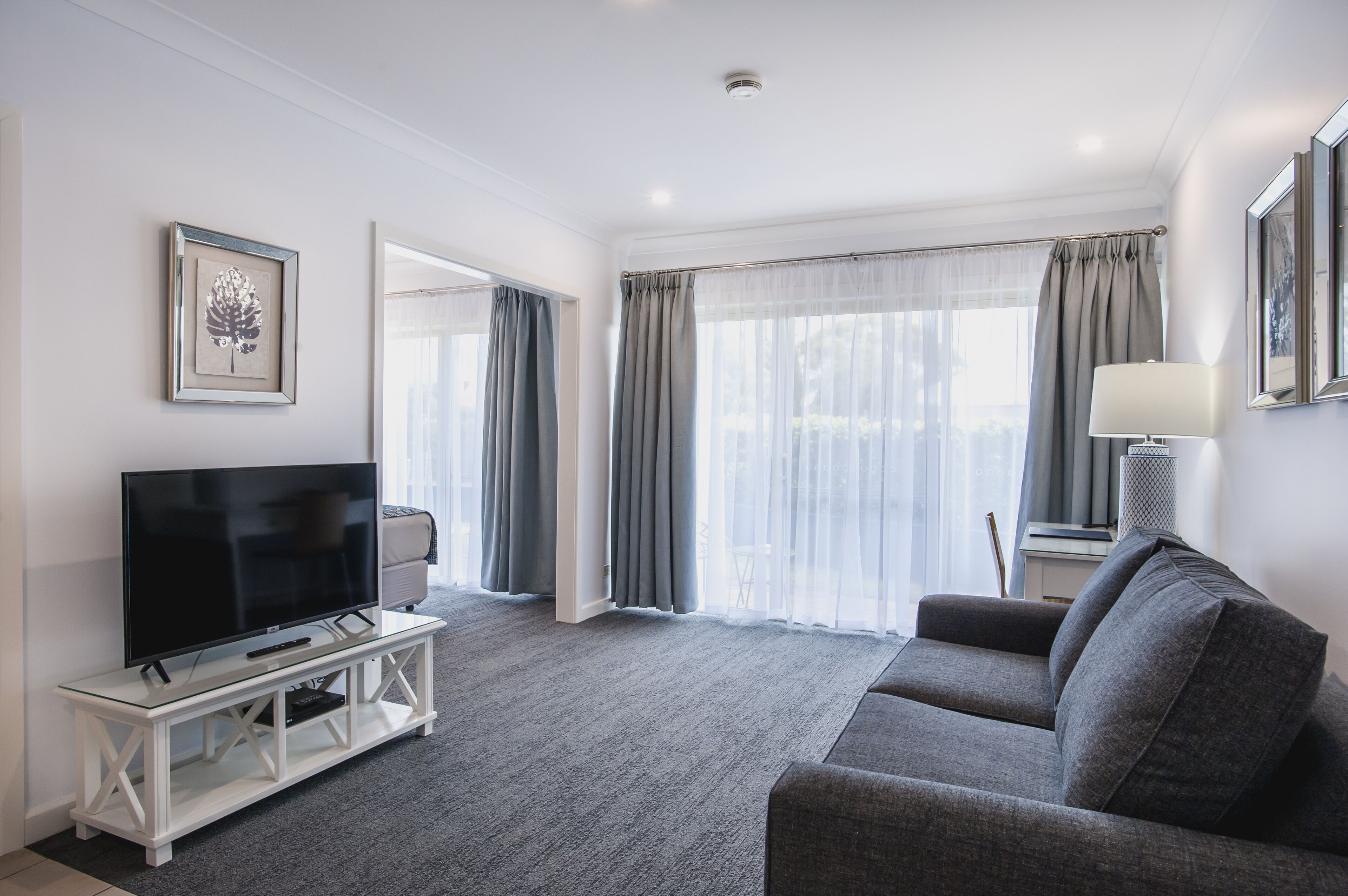 A modern living room with a flat-screen TV on a white console, a grey sofa, and a large window with grey curtains.
