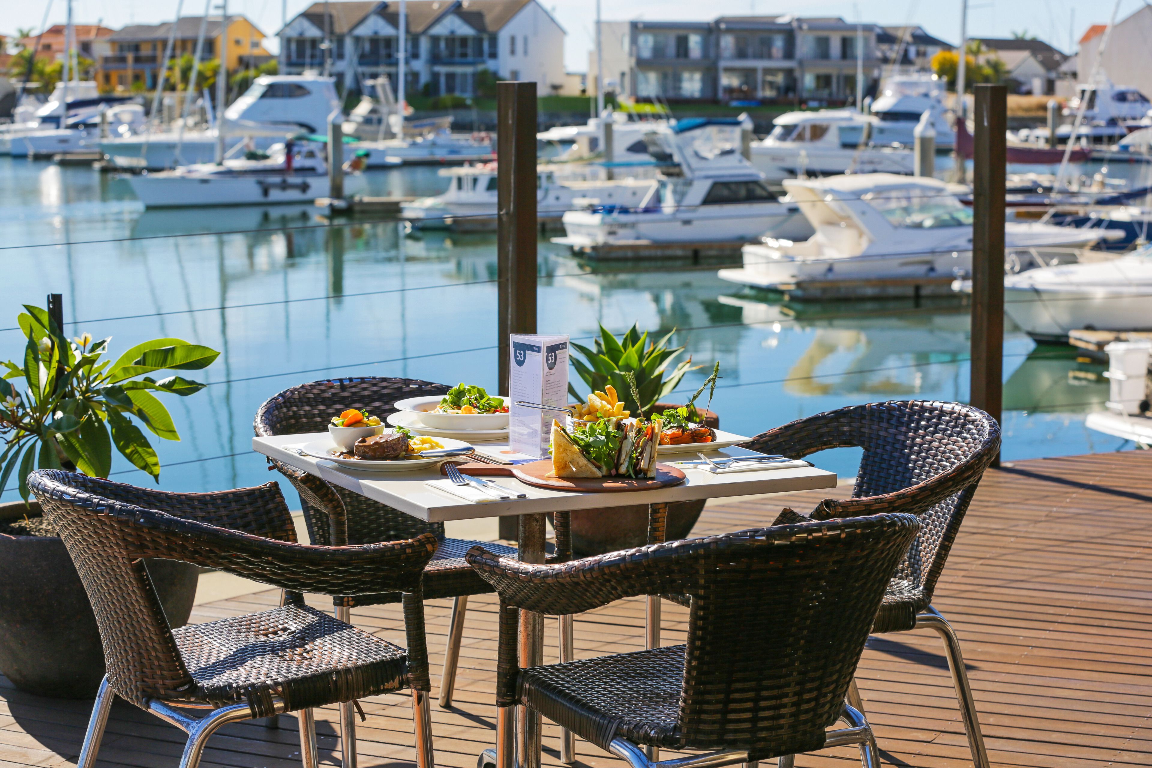Mesa con sillas y un menú en una terraza de madera con vista a un puerto deportivo y barcos.