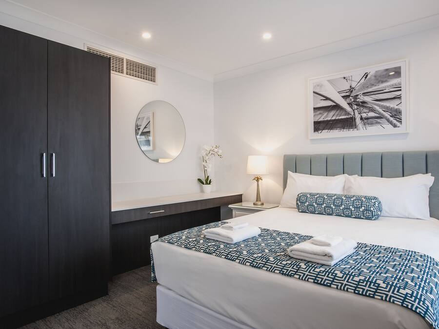 A modern hotel room with a large bed, dark wooden wardrobe, a round mirror, a bedside table with a lamp, and a framed artwork on the wall.