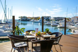 Mesa con sillas y un menú en una terraza de madera con vista a un puerto deportivo y barcos.