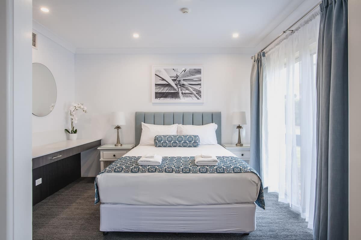 A modern hotel room with a large bed, a bedside table with a lamp, a wall-mounted mirror, and a window with curtains.