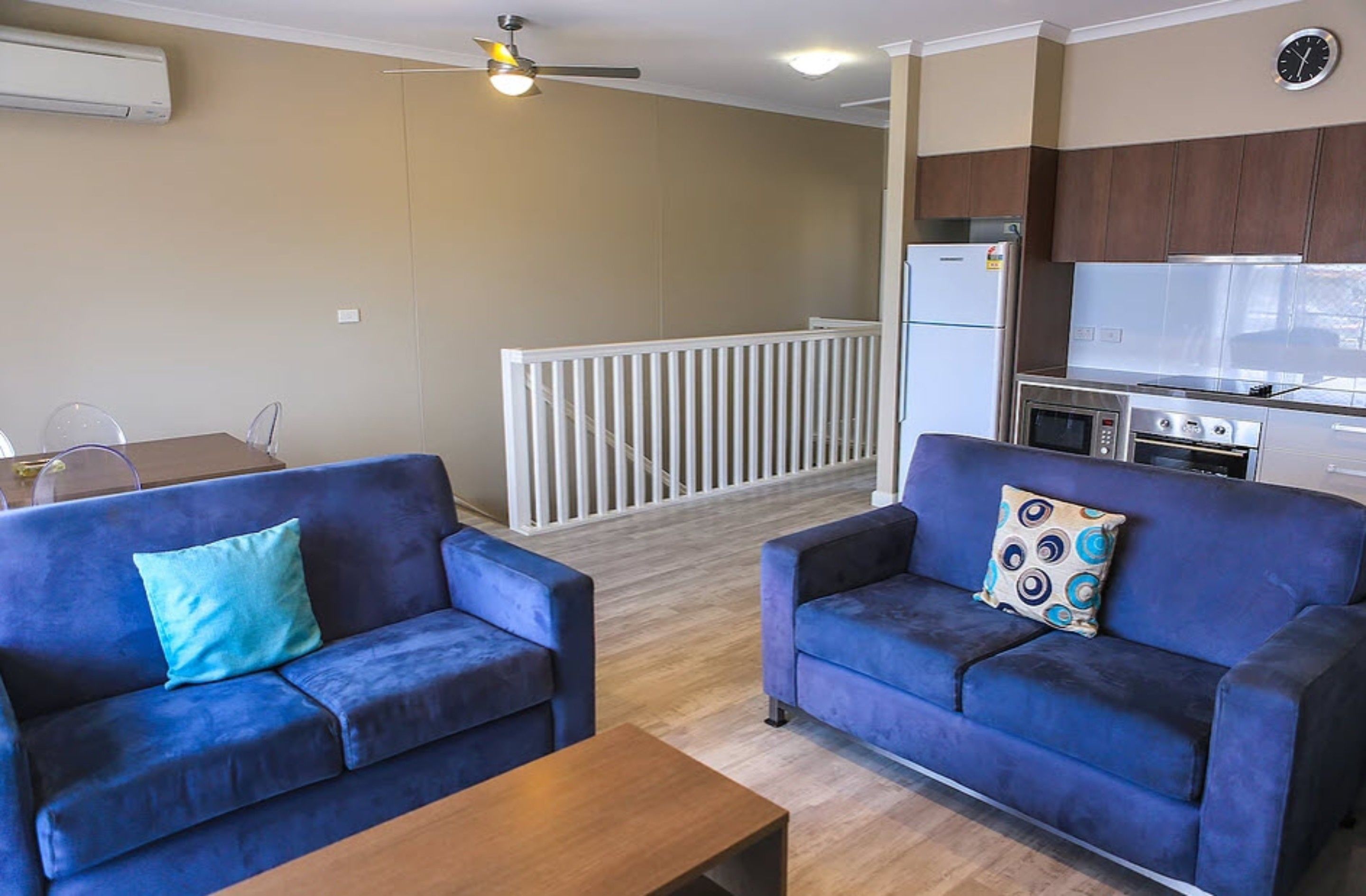 A living room with two blue sofas, a wooden coffee table, and a dining area with a table and chairs. A kitchen with modern appliances is visible in the background.