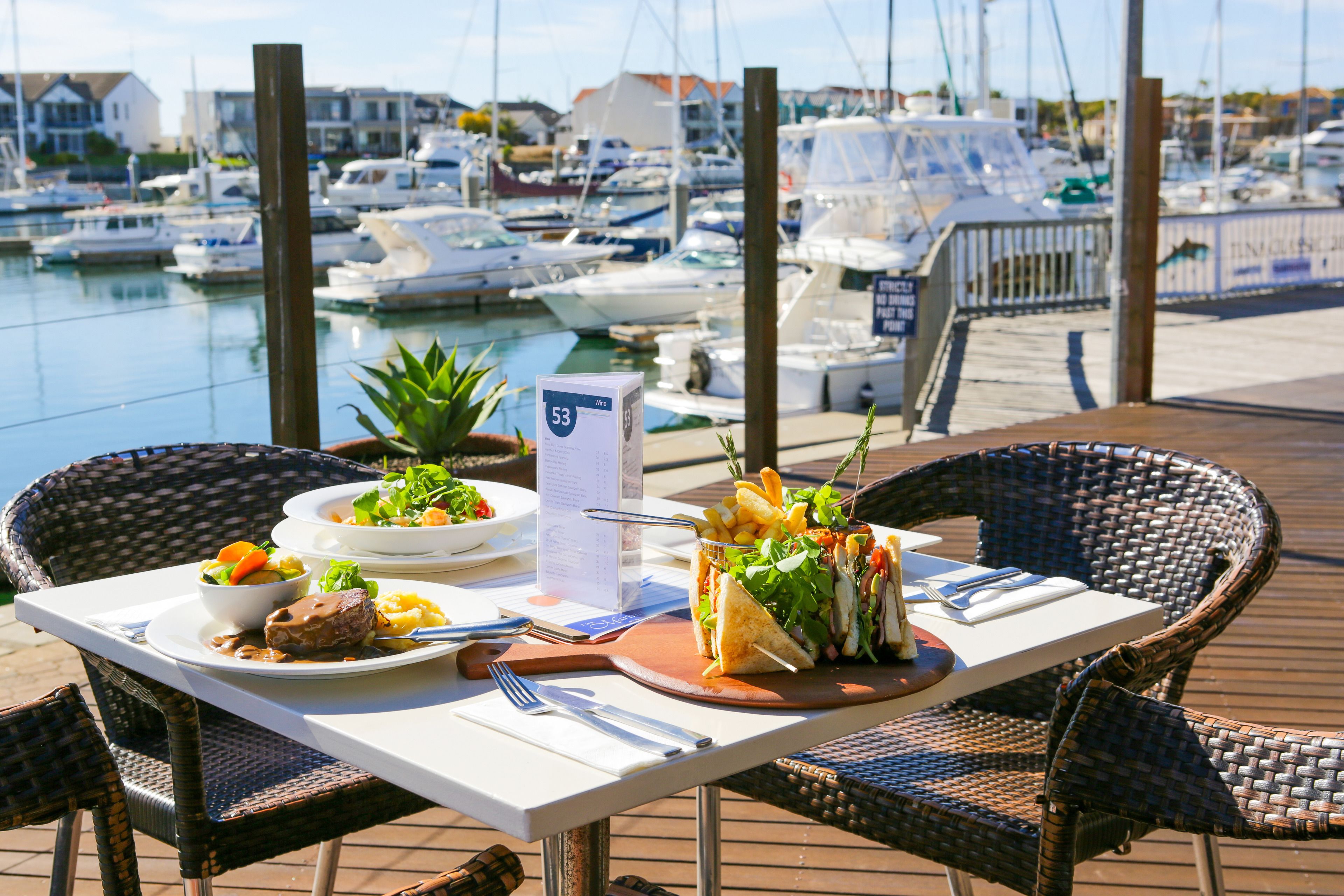 Una mesa de comedor con comida, sillas y vista a barcos en un puerto deportivo.