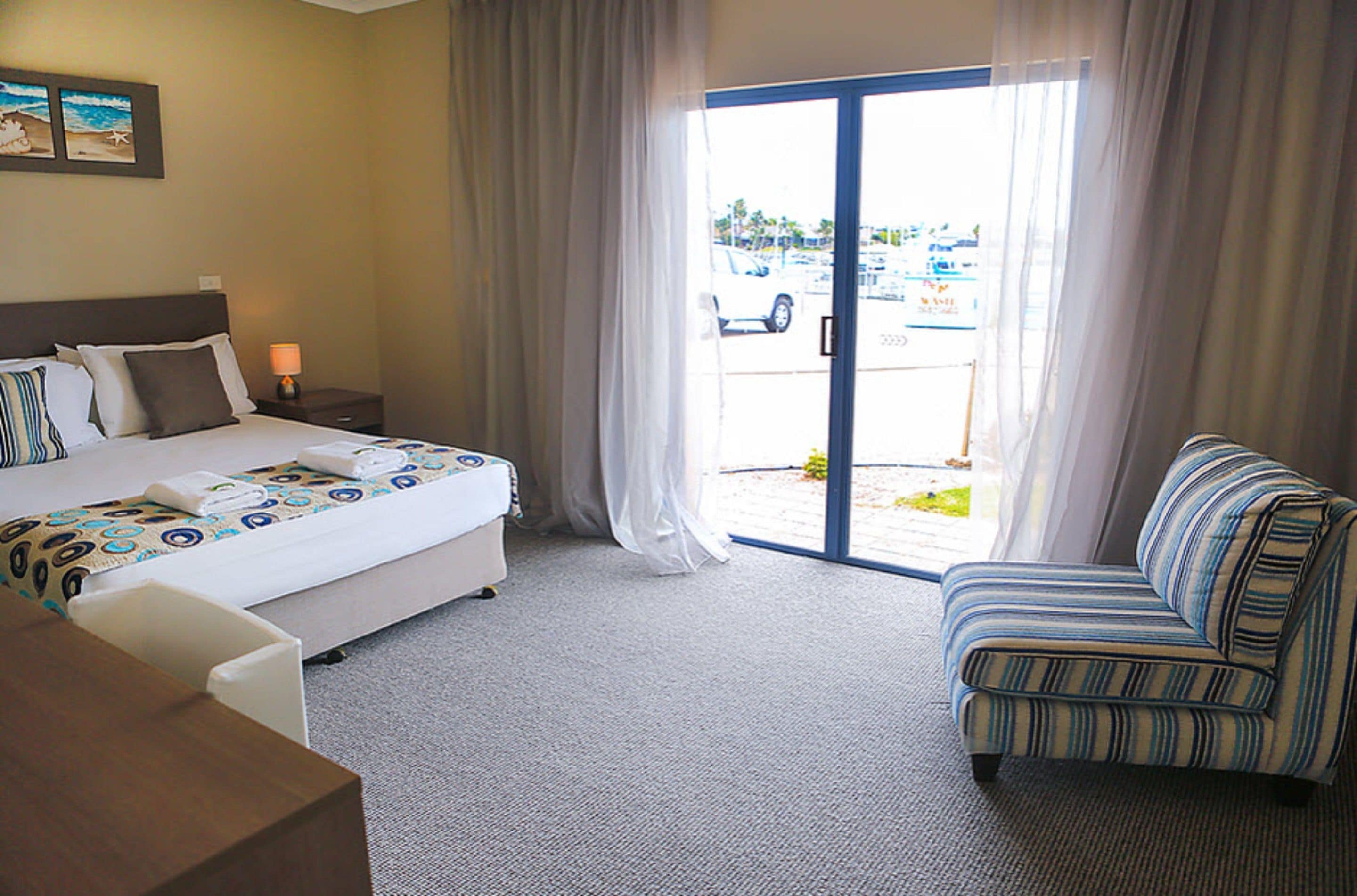 A hotel room with a bed, a desk, a chair, and a view of the parking lot through the glass door.