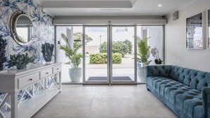 A modern hotel lobby with a white reception desk, a blue sofa, and large glass doors leading to an outdoor area with plants and a view of the city. - The Marina Hotel (Port Lincoln)