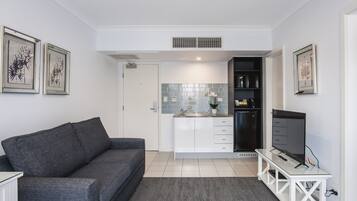 Una sala de estar moderna con un sofá gris, un televisor sobre un mueble blanco y una zona de cocina con un lavaplatos, armarios y un refrigerador.