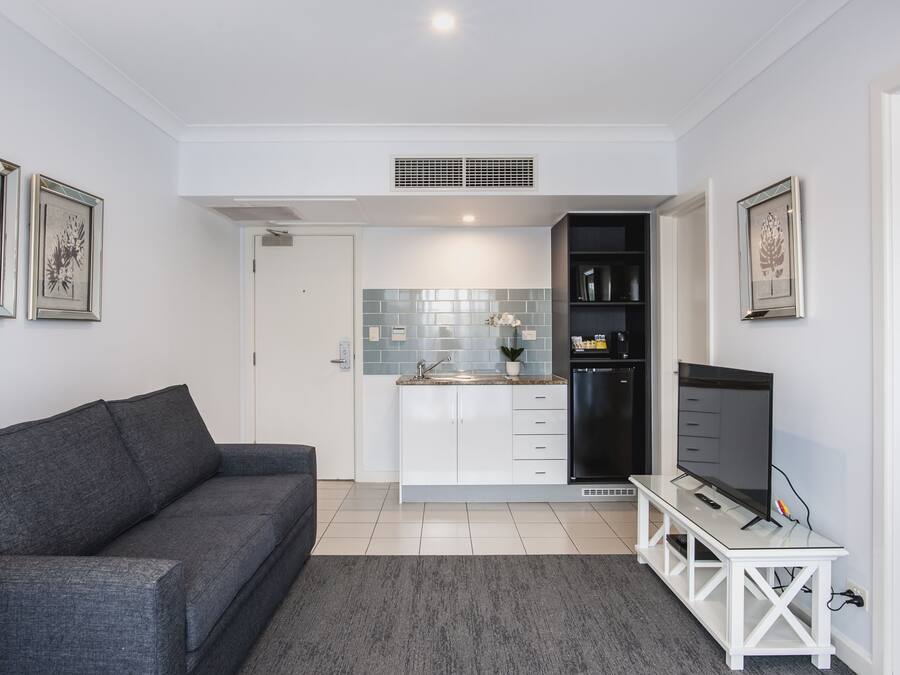 Una sala de estar moderna con un sofá gris, un televisor sobre un mueble blanco y una zona de cocina con un lavaplatos, armarios y un refrigerador.