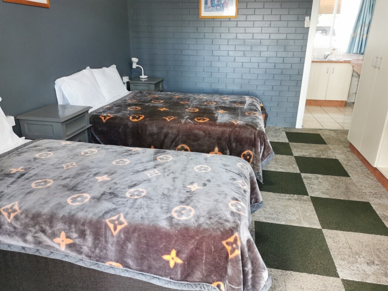Ein Hotelzimmer mit zwei Betten, einem kleinen Schreibtisch, einem Nachttisch, einem Fenster mit Vorhängen und einem Schachbrettfußboden.