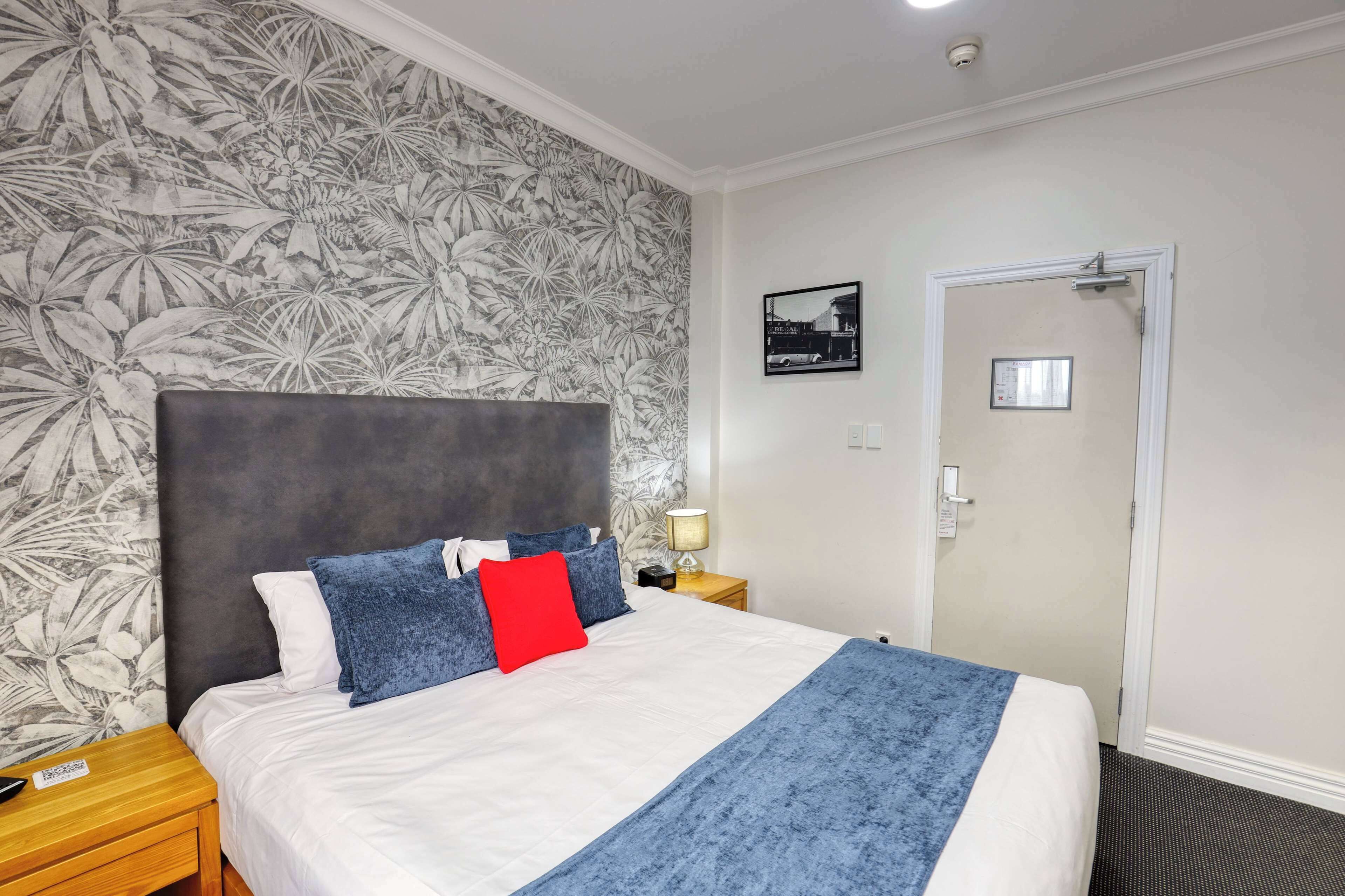 Habitación de hotel con una cama grande, cabecero, mesita de noche, lámpara, puerta y pared con estampado de hojas tropicales.