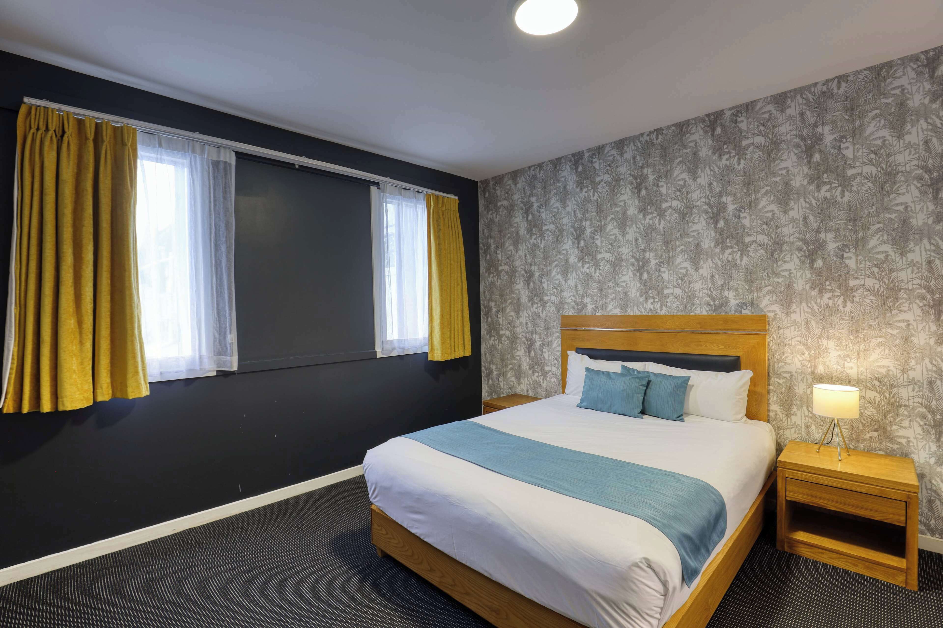 Une chambre d’hôtel avec un grand lit, une table de chevet avec une lampe, une fenêtre avec des rideaux jaunes et un papier peint à motifs.