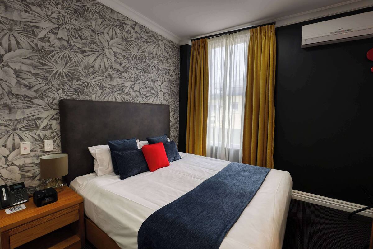 A hotel room with a bed, a nightstand with a lamp, a telephone, and a wall-mounted air conditioner.