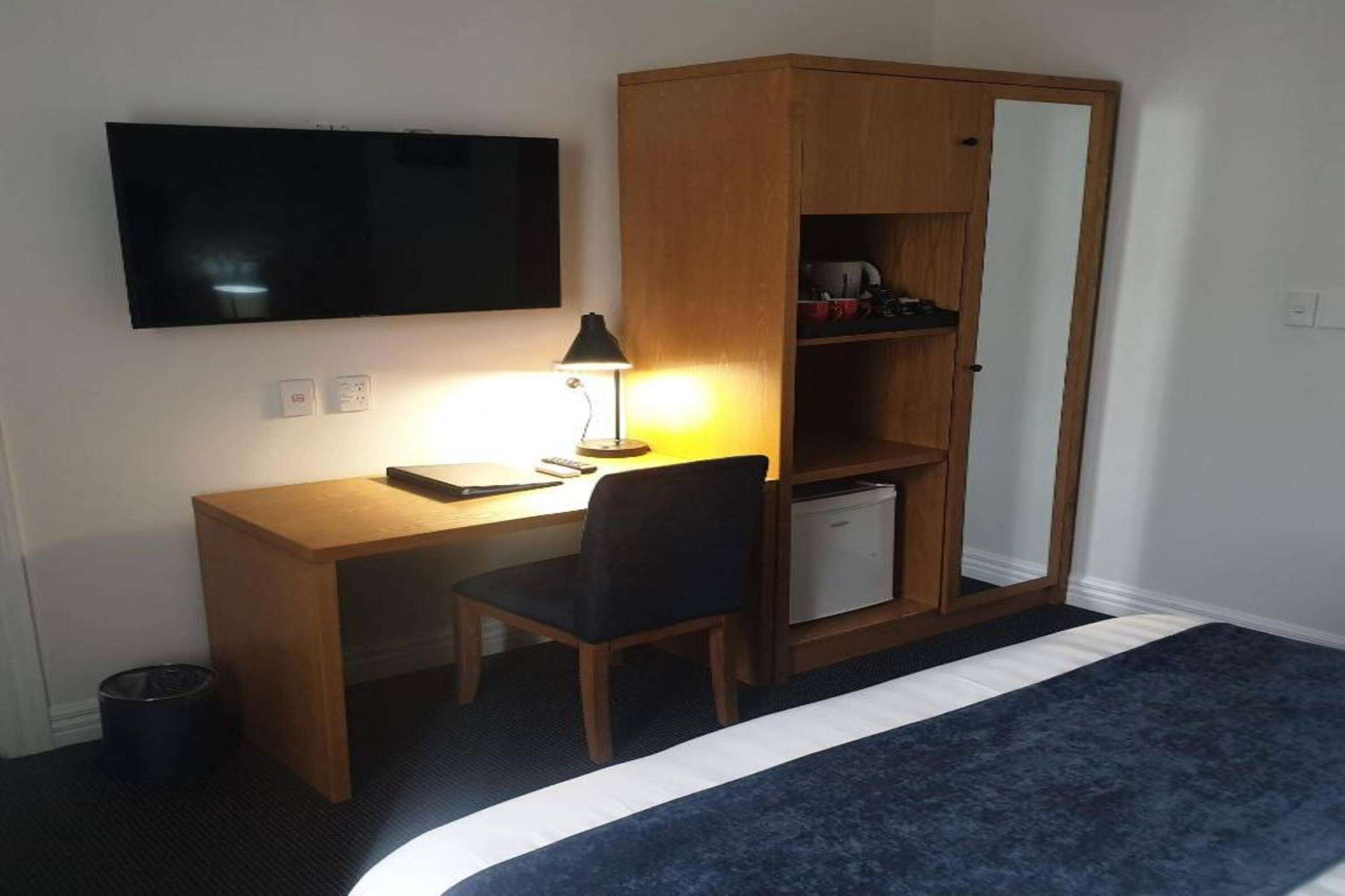A hotel room with a wooden desk, a chair, a bed with a blue and white bedspread, a television mounted on the wall, and a wardrobe with a mirror.