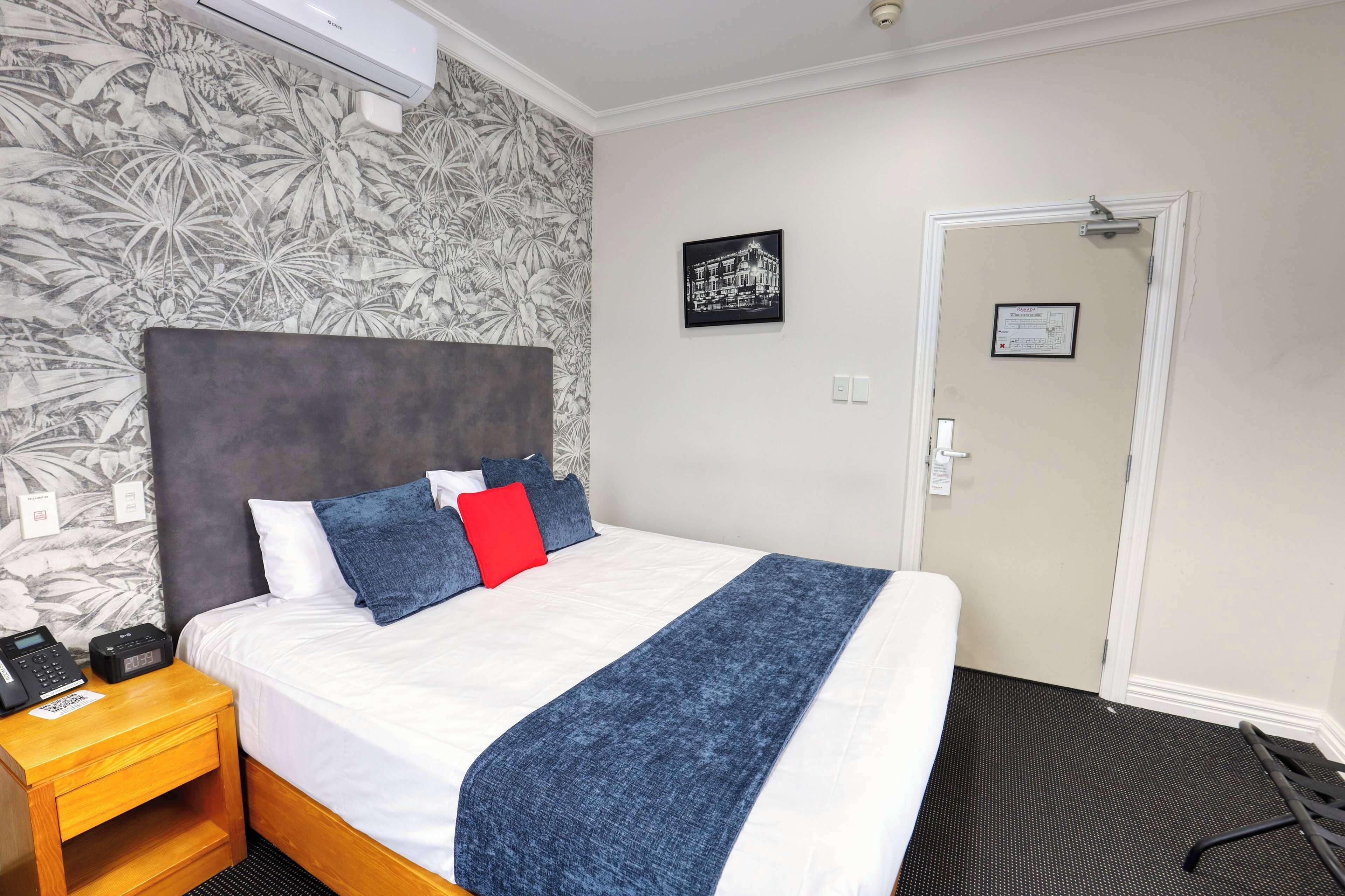 Habitación de hotel con cama, mesita de noche con teléfono y radio, una puerta y pared con papel tapiz tropical.