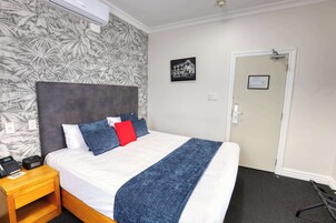 Habitación de hotel con cama, mesita de noche con teléfono y radio, una puerta y pared con papel tapiz tropical.