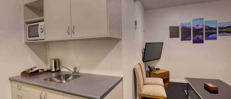 Cocina compacta con microondas, lavamanos y armario. Hay una mesa pequeña con una silla y un televisor de pantalla plana en la pared.