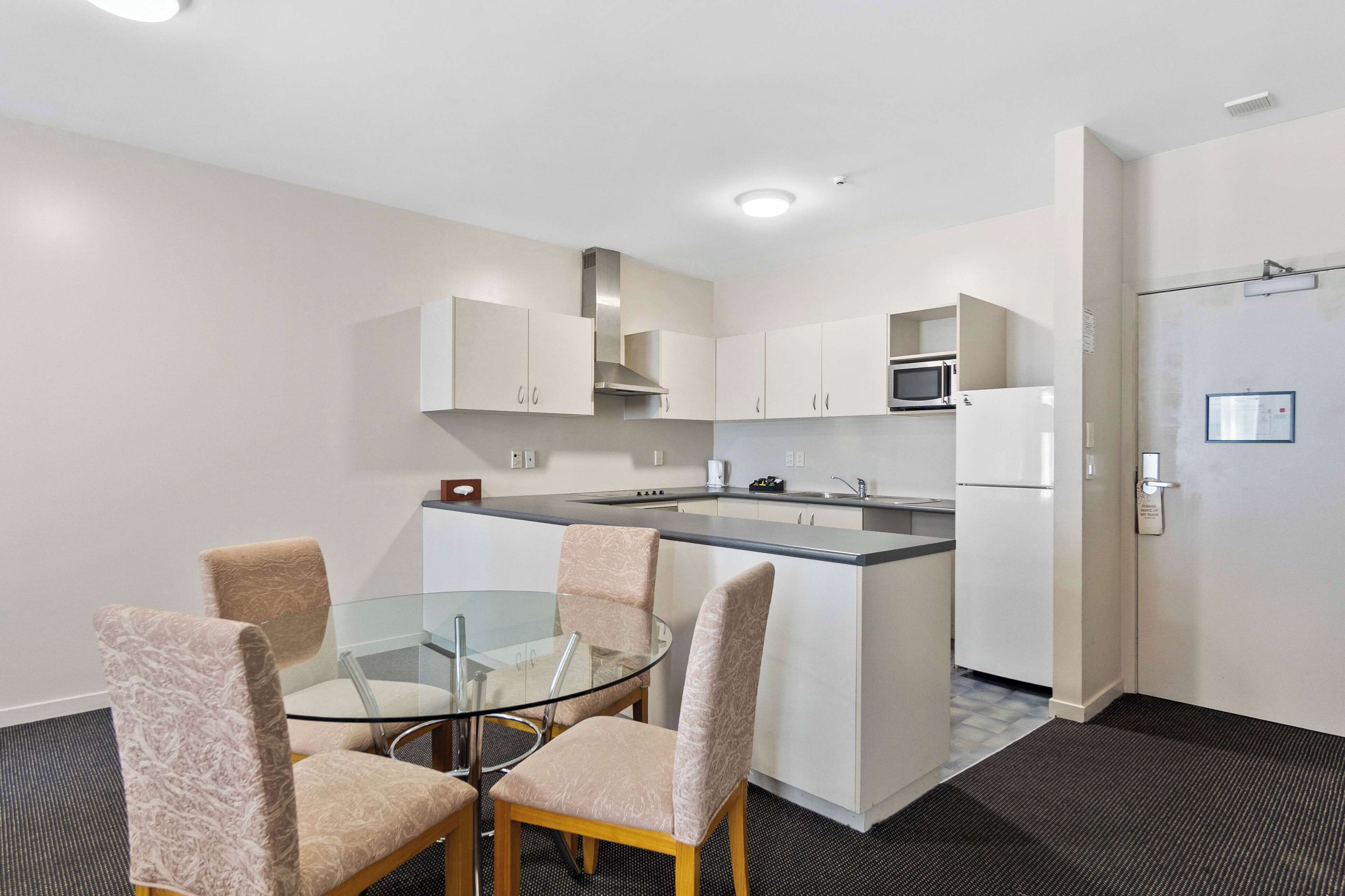 A modern kitchen with white cabinets, a stainless steel range hood, and a microwave. There is a glass dining table with four chairs.