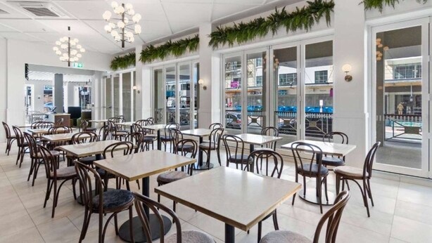 A spacious indoor dining area with multiple tables and chairs, large windows, and decorative greenery. - Ramada by Wyndham Hamilton City Center (Hamilton)