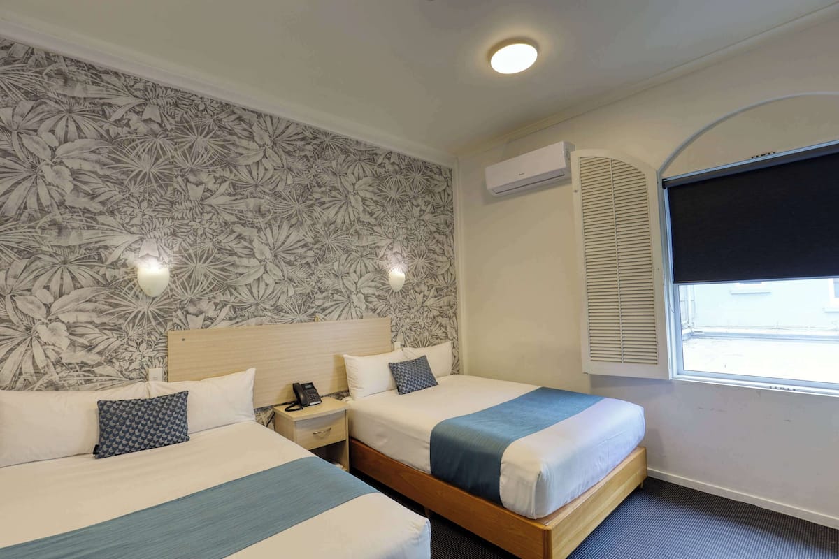A hotel room with two beds, a floral wallpaper, an air conditioning unit, and a window with blinds.