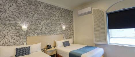 A hotel room with two beds, a floral wallpaper, an air conditioning unit, and a window with blinds.