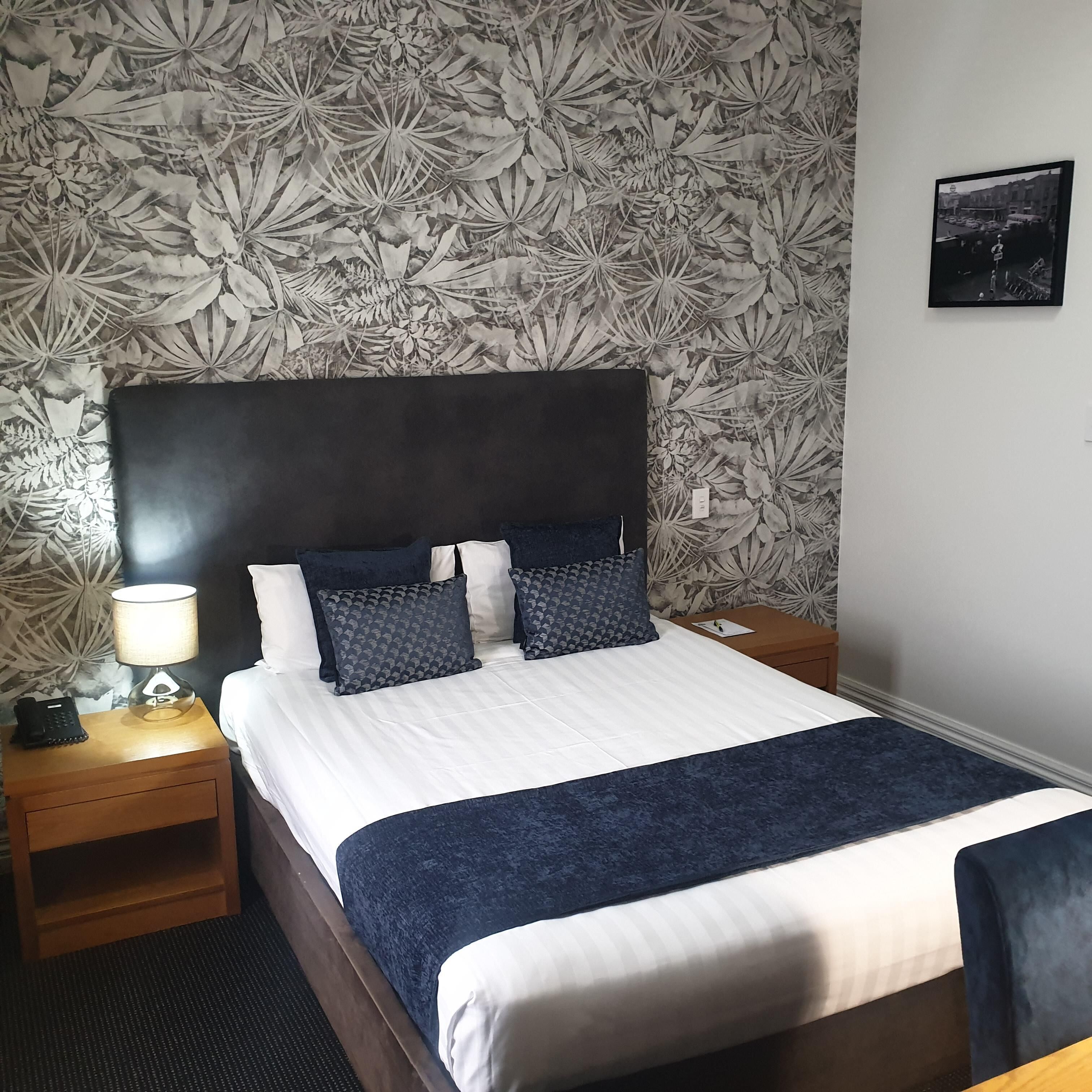 A neatly made bed with a dark headboard, white bedding, and a blue accent throw. There is a nightstand with a lamp and a phone, and a framed picture on the wall.