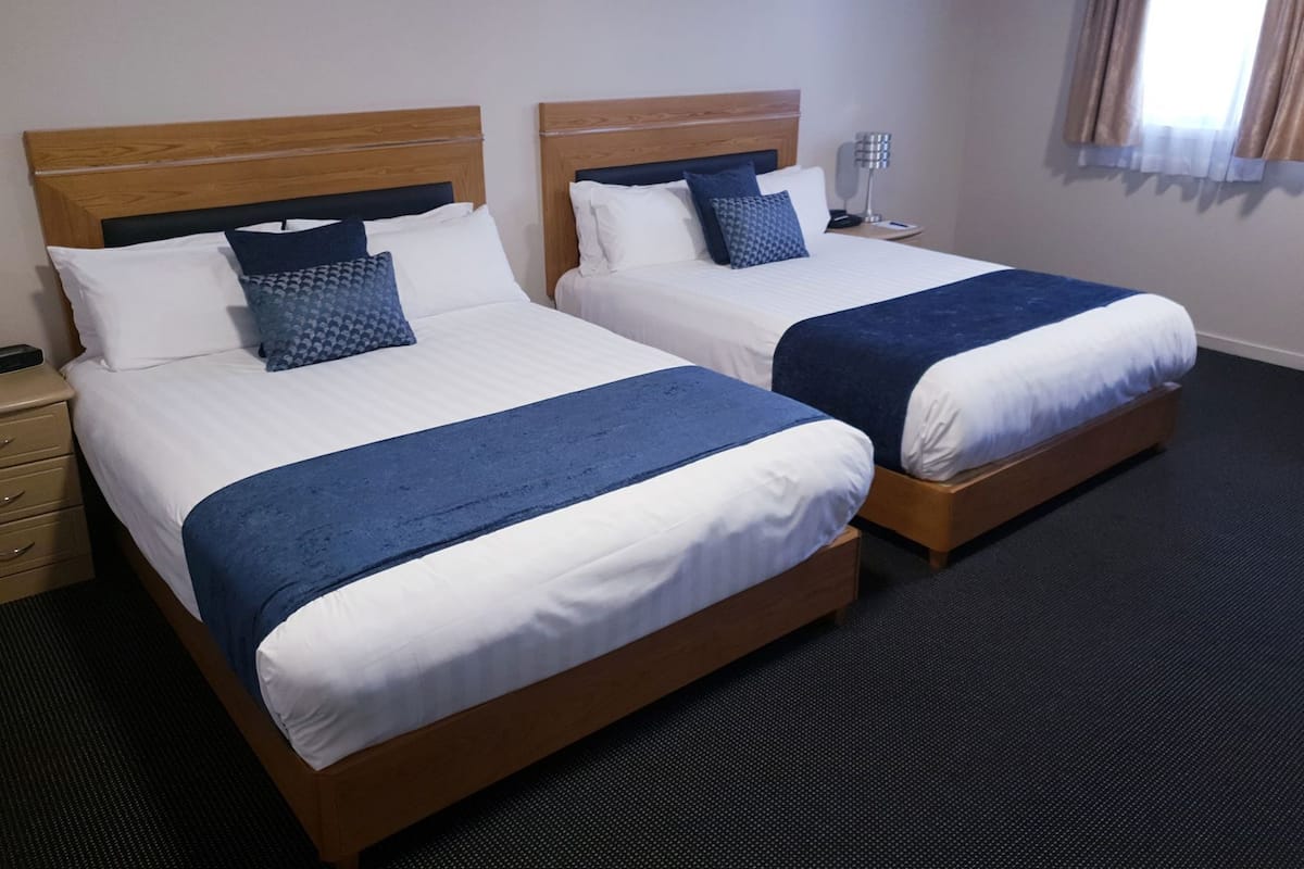 A hotel room with two beds, each with a wooden headboard, white bedding, and a blue accent pillow. There is a bedside table with a lamp, and the room has a carpeted floor.