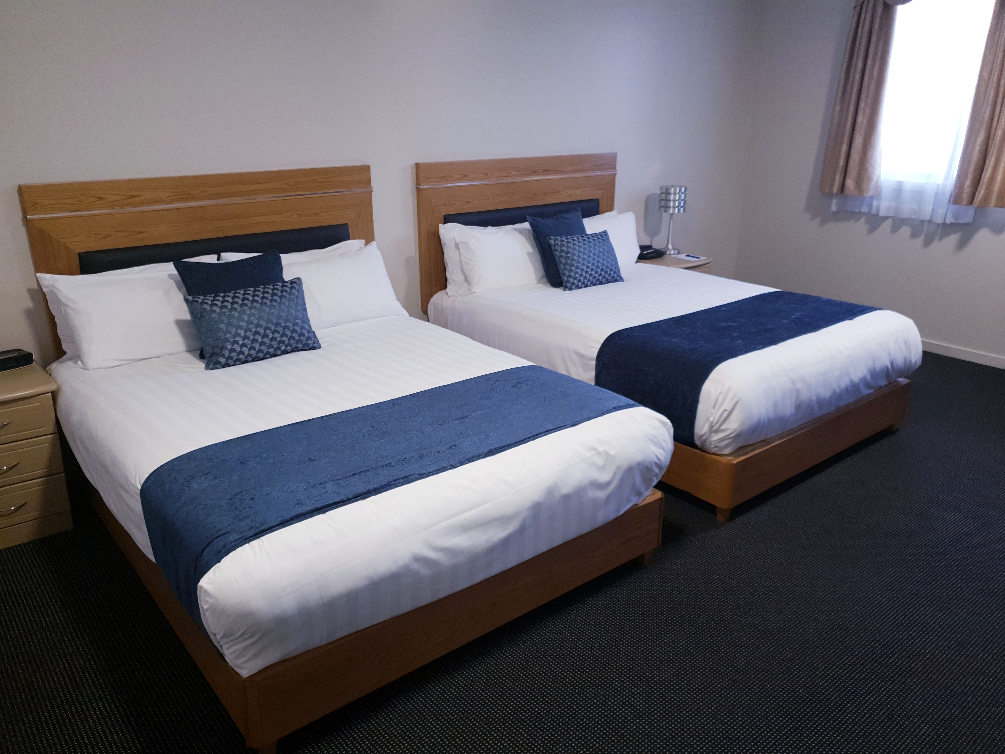 Habitación de hotel con dos camas, cada una con cabecero de madera, ropa de cama blanca y mantas azules. Se ve un mesita de noche con una lámpara y un teléfono.