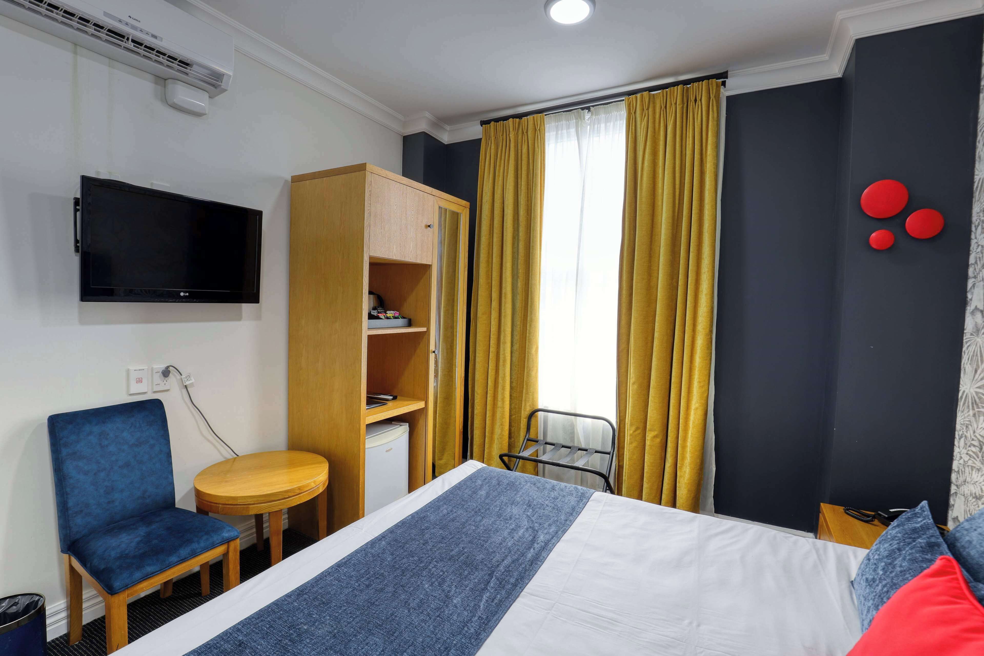 A hotel room with a bed, a blue chair, a small table, a wardrobe, a TV, and a window with yellow curtains.