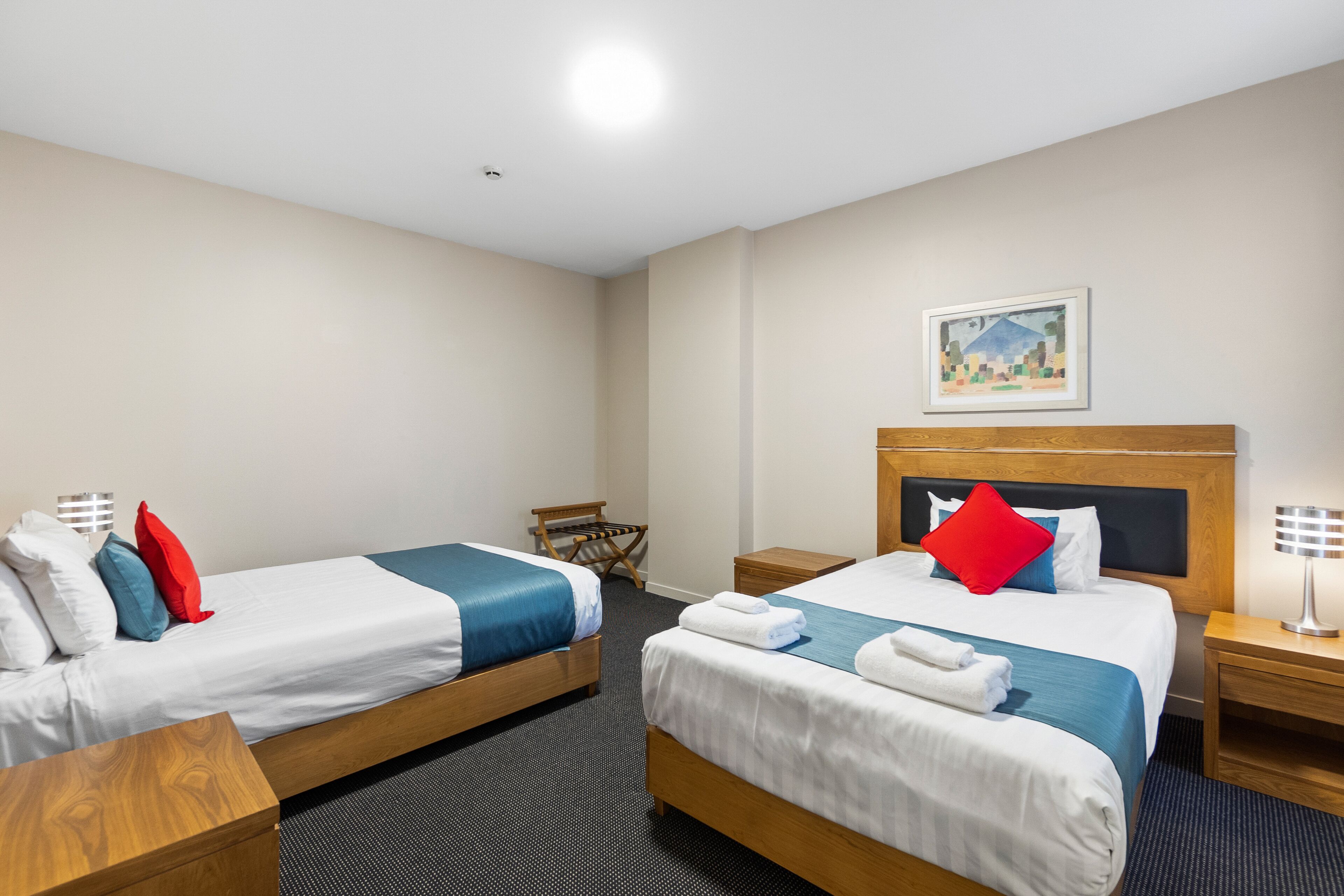 A hotel room with two beds, a wooden headboard, a small table, a chair, and a nightstand with a lamp.