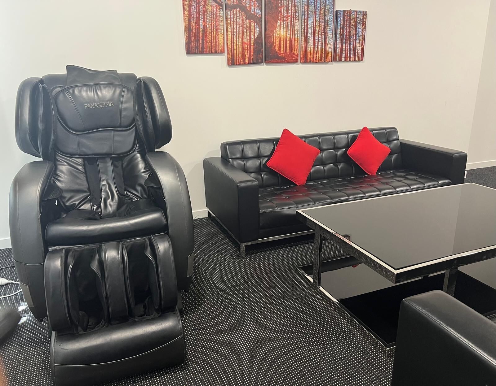 Una silla de masaje, un sofá de cuero negro con cojines rojos y una mesa de centro negra en una habitación con suelo moteado.