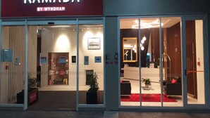 A hotel entrance with a 'RAMADA BY WYNDHAM' sign, glass doors, a reception desk, and a red carpet. - Ramada by Wyndham Hamilton City Center (Hamilton)