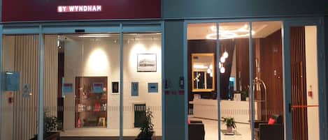 A hotel entrance with a 'RAMADA BY WYNDHAM' sign, glass doors, a reception desk, and a red carpet.