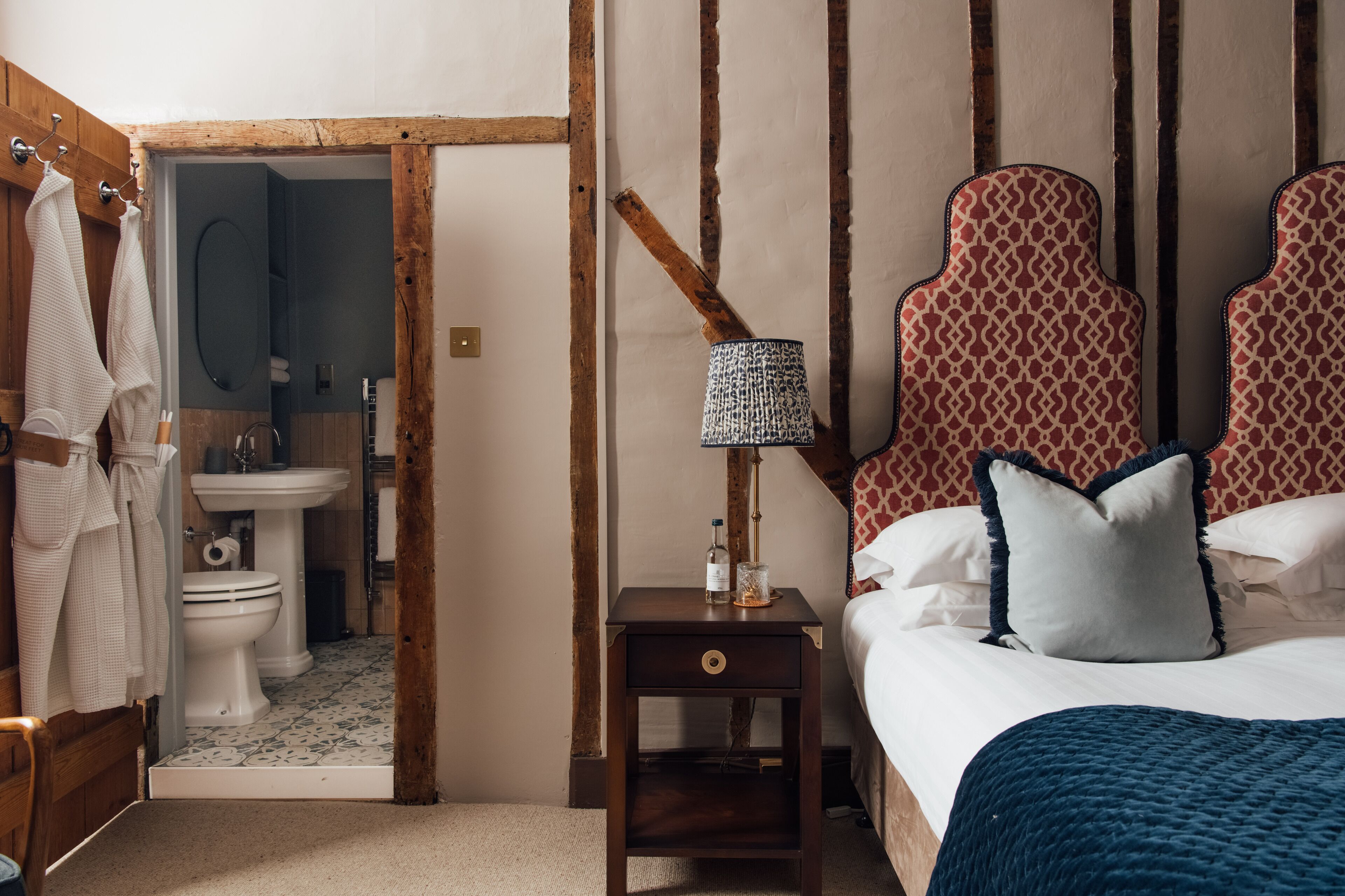 A bedroom with a wooden headboard, a bedside table with a lamp, a bed with pillows, and a view into the bathroom.